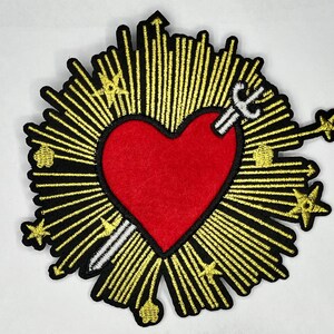 May include: A red heart with a white sword piercing through it, surrounded by a golden halo of rays. The heart is embroidered on a black fabric.