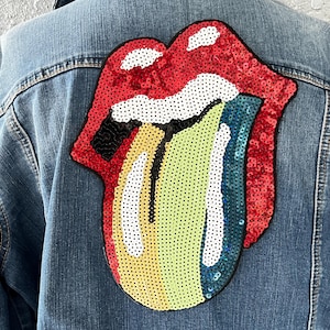 May include: A denim jacket with a large, sequined Rolling Stones tongue logo on the back. The tongue is red, yellow, blue, and white, with black outlining. The jacket is a light blue wash.