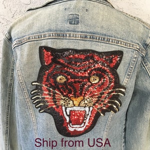 May include: Light blue denim jacket with a large, sequined tiger patch on the back. The tiger design features red, black, and gold sequins, with an open mouth and visible teeth. The jacket has a classic collar and button closures.