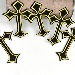 May include: A collection of black cross patches with gold trim. The crosses have a gothic design, with a diamond shape at the top and bottom of each arm. The patches are likely for clothing or accessories.