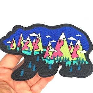 May include: An embroidered iron-on patch featuring a black bear silhouette with a colorful mountain range scene inside. The mountains are pink, green, and yellow with white snow peaks. The background is a dark blue sky with white clouds.