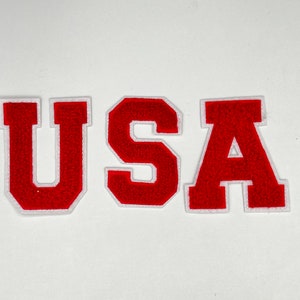 Może przedstawiać: Czerwone filcowe litery układające się w napis "USA" z białymi obramowaniami. Litery znajdują się na białym tle.
