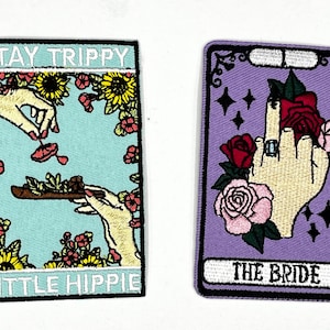 May include: Two embroidered patches with a floral theme. The first patch is blue with white text that reads "Stay Trippy Little Hippie". The second patch is purple with white text that reads "The Bride".