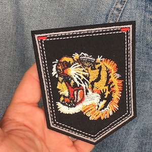 May include: A black embroidered patch with a detailed image of a tiger's head with orange, yellow, white, and red stitching. The patch is sewn onto a denim jacket.