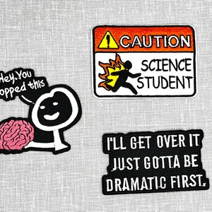 May include: Three embroidered iron-on patches. The first patch is a cartoon of a stick figure holding a pink brain with the text "Hey. You dropped this". The second patch is a yellow and orange caution sign with the text "CAUTION SCIENCE STUDENT". The third patch is black with white text that reads "I'LL GET OVER IT JUST GOTTA BE DRAMATIC FIRST."