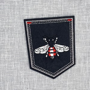 May include: A black embroidered patch with a white bee with red stripes on a gray background.