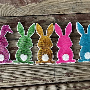 May include: Six colorful felt bunny rabbit shapes with white felt tails. The bunnies are pink, green, gold, pink, blue, and blue. The bunnies are all facing the same direction.