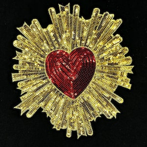 May include: A heart-shaped sequin patch with a red center and gold rays radiating outwards. The patch is embellished with gold sequins, creating a sparkling effect. The design is set against a black background.