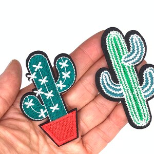 May include: Two embroidered cactus patches. One cactus is green with white flowers and is in a red pot. The other cactus is green and blue with white accents.