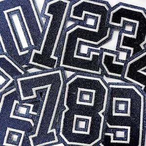 May include: A collection of navy blue embroidered number patches with white borders. The numbers are in a classic varsity style, with a textured denim-like appearance. The patches are ideal for customizing apparel or accessories.