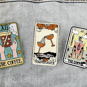 May include: Three embroidered tarot card patches featuring the Temperance, The Coffee, and The Coffee cards. The Temperance card features two golden chalices pouring liquid into each other. The Coffee cards feature a hand holding a cup of coffee.