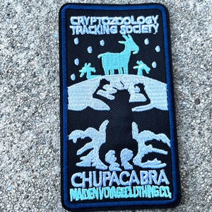 May include: A blue and white embroidered patch with the text "Cryptozoology Tracking Society" and "Chupacabra Maiden Voyage Clothing Co.". The patch features a blue silhouette of a goat with a black silhouette of a creature behind it.