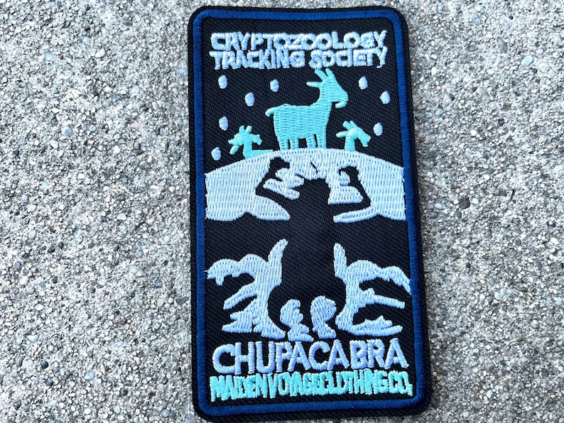 May include: A blue and white embroidered patch with the text "Cryptozoology Tracking Society" and "Chupacabra Maiden Voyage Clothing Co.". The patch features a blue silhouette of a goat with a black silhouette of a creature behind it.