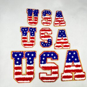 May include: Three sets of the letters "USA" in a stacked arrangement. Each letter is made of red, white, and blue fabric with a gold outline and a fuzzy texture. The letters are arranged in a pyramid shape, with the largest set at the bottom and the smallest set at the top.