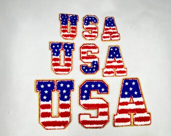 May include: Three sets of the letters "USA" in a stacked arrangement. Each letter is made of red, white, and blue fabric with a gold outline and a fuzzy texture. The letters are arranged in a pyramid shape, with the largest set at the bottom and the smallest set at the top.