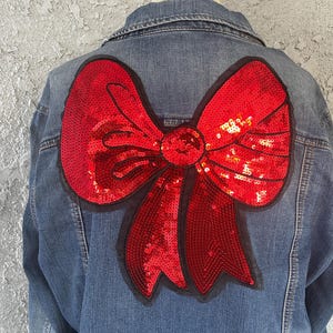 May include: A denim jacket with a large, red sequined bow on the back. The bow is outlined in black and is the focal point of the jacket. The jacket is a medium wash denim.