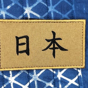 May include: A rectangular patch with the Japanese characters for "Japan" embroidered in black on a tan background. The patch is set against a blue fabric with a white geometric pattern.