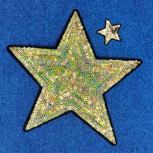 May include: Two star-shaped patches on a blue denim background. The larger star is filled with gold and iridescent sequins and outlined in black. A smaller star, also sequined, is above and to the right of the larger star.