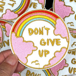 May include: Embroidered patch with a rainbow design and the words "DON'T GIVE UP" in gold lettering. The patch features pink clouds and a gold border. The patch is round and has a white background.