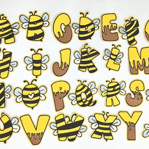 May include: A collection of embroidered alphabet patches, each designed with a bee theme. The letters are yellow with black stripes, resembling bees, and some have brown accents. The patches are ideal for decorating clothing or accessories.