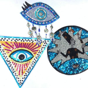 May include: Three embroidered patches with blue and silver sequins. One patch is a triangle with a blue eye inside. Another patch is a circle with a blue eye inside. The third patch is a circle with a black figure with stars inside.