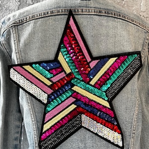 May include: A light-wash denim jacket with a large, colorful star patch on the back. The star is filled with sequins in various colors, including pink, teal, red, and gold, and is outlined in black.