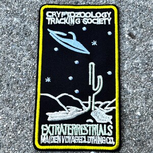 May include: A black and yellow embroidered patch with a white UFO, cactus, and stars. The text reads "Cryptozoology Tracking Society" and "Extraterrestrials Maiden Voyage Clothing Co."