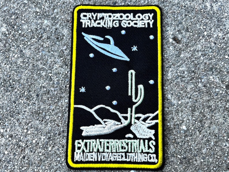 May include: A black and yellow embroidered patch with a white UFO, cactus, and stars. The text reads "Cryptozoology Tracking Society" and "Extraterrestrials Maiden Voyage Clothing Co."
