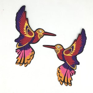 May include: Two embroidered hummingbird patches in shades of purple, red, orange, and yellow. The hummingbirds are flying with their wings spread out.