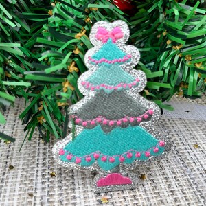 May include: A silver glitter Christmas tree applique with a pink bow, turquoise and gray layers, and pink lights.