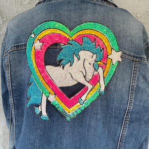 May include: A denim jacket with a sequined heart-shaped patch featuring a white unicorn with a blue mane and tail. The heart is outlined in pink, yellow, and green, with white star accents. The jacket is a medium wash blue.