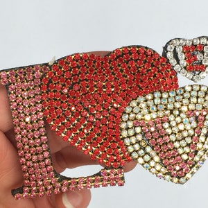 May include: A red, pink, and clear rhinestone applique in the shape of the word "I LOVE" with a small heart at the end.