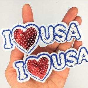 May include: Two white fabric patches with blue trim and red sequin hearts. The patches read "I love USA".