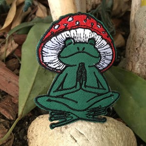 May include: Embroidered patch featuring a green frog in a meditative pose, wearing a red mushroom cap with white spots. The frog is sitting cross-legged with hands together. The patch is on a stone.