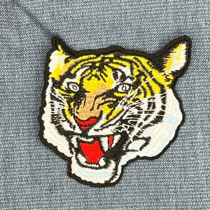 May include: A black and white embroidered patch of a tiger's face with yellow stripes and a red mouth.