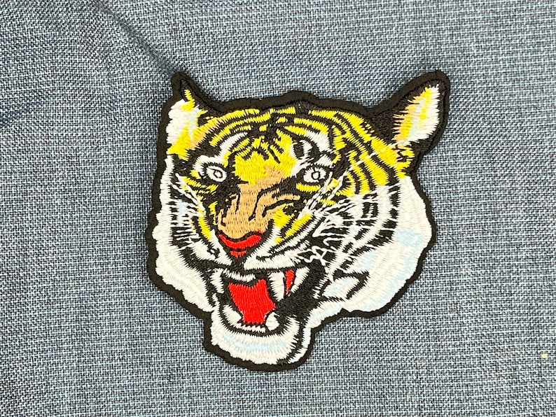 May include: A black and white embroidered patch of a tiger's face with yellow stripes and a red mouth.