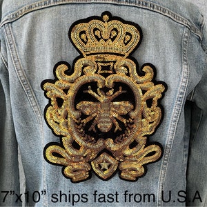 May include: A gold sequined iron-on patch with a crown, ornate scrollwork, and a bee in the center. The patch is 7 inches by 10 inches.