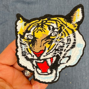 May include: A close-up of an embroidered iron-on patch featuring a tiger's face with yellow and white stripes, black outlines, and a red open mouth.