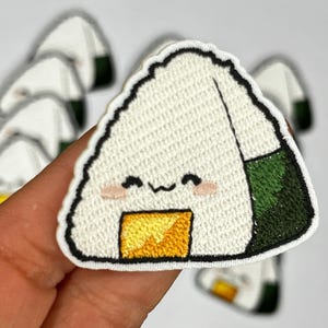 May include: A white embroidered patch of a cartoon onigiri, a Japanese rice ball, with a smiling face and a yellow filling. The onigiri has a green seaweed wrap.