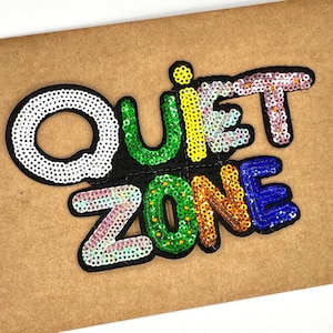 May include: A colorful sequined patch that reads "Quiet Zone" in rainbow colors. The letters are outlined in black and the background is a brown fabric.