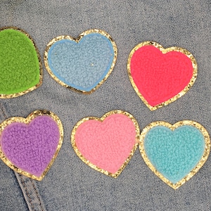 May include: Six heart-shaped patches with gold glitter trim. The hearts are made of fuzzy fabric and are in the colors green, light blue, pink, red, purple, and light blue.
