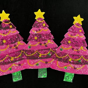 May include: Three pink Christmas tree ornaments with yellow star toppers. The trees are decorated with sequins and beads, featuring yellow and green accents. The trees have green beaded bases and are set against a black background.