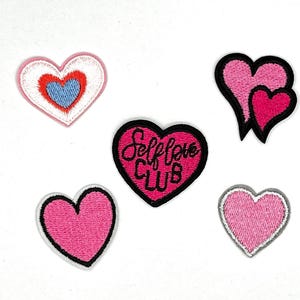 May include: Five embroidered iron-on patches in the shape of hearts. One heart is pink with white outline and the text "Self Love Club" in black. Two hearts are pink with white outlines. One heart is pink with a white outline and a smaller blue heart inside. One heart is pink with a white outline and a smaller pink heart inside.