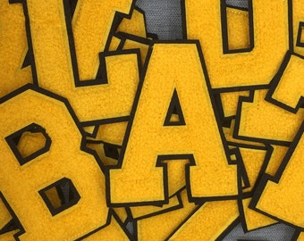May include: A close-up of yellow felt letters with black outlines. The letters are stacked on top of each other and spell out the word "BACK".