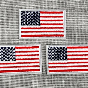 May include: Three small embroidered American flag patches with white stars on a blue field and red and white stripes. The patches are on a gray textured background.