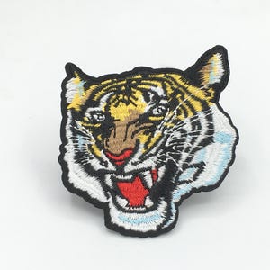 May include: Embroidered iron-on patch featuring a tiger's face with yellow, orange, white, and black fur. The tiger has a red mouth and white teeth.