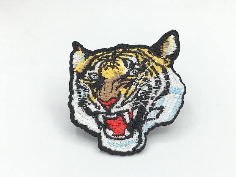 May include: Embroidered iron-on patch featuring a tiger's face with yellow, orange, white, and black fur. The tiger has a red mouth and white teeth.