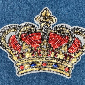 May include: A gold and red sequined crown with a pointed top. The crown is on a blue denim background.