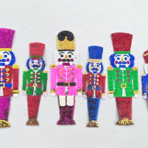 May include: Six colorful nutcracker ornaments with beaded details. The ornaments are made of fabric and sequins and feature different colors and designs.