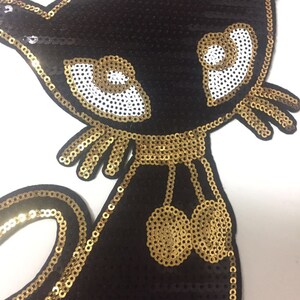 May include: A black and gold sequined cat applique with large white eyes and gold embellishments. The cat has a gold collar and two gold balls hanging from it.